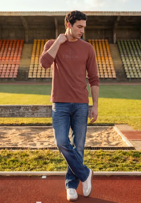 Longsleeve met tonale print raw copper red Ein Mann in Jeans und langärmeligem T-Shirt steht entspannt vor Stadiontribüne.