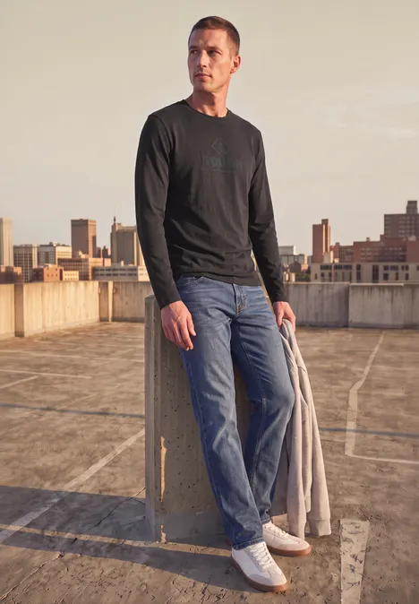 T-shirt à manches longues avec imprimé ton sur ton shadow brown Mann auf Parkdeck, trägt schwarzen Pullover, Jeans, Pullover in der Hand, Skyline im Hintergrund.