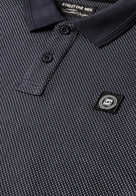 Nahaufnahme eines schwarzen Poloshirts mit feinem Muster, Kragen und einem markanten S1-Logo-Pin.