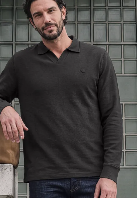 Ein Mann lehnt an einer Wand aus Glasbausteinen, trägt ein schwarzes Langarm-Polo und Jeans.