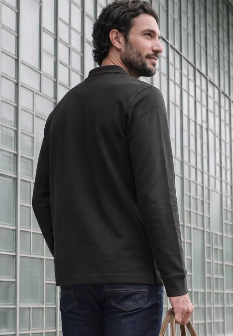 Ein bärtiger Mann steht in schwarzem Hemd und Jeans vor einer Glasblockwand und schaut seitlich nach rechts.