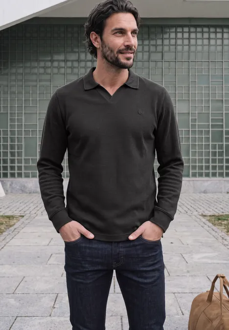 Ein Mann steht lässig in dunkler Polo langärmelig, Hände in Taschen, Tasche neben ihm auf dem Gehweg.