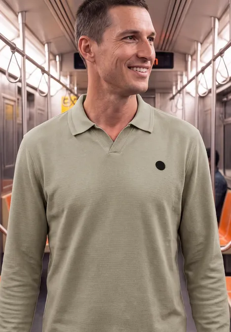 Ein lächelnder Mann in einer U-Bahn trägt ein olivgrünes Polohemd mit rundem Logo auf der Brust.