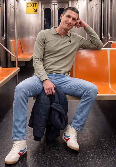 Ein Mann sitzt in einer leeren U-Bahn, trägt einen beigen Pullover, Jeans und hält eine Jacke.