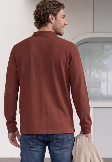 Ein Mann steht von hinten in einem braunen Langarmshirt und hält locker eine Jacke.
