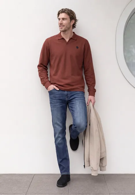 Mann in rotem Langarm-Polo, Jeans und schwarzen Schuhen lehnt an weißer Wand und hält helle Jacke.