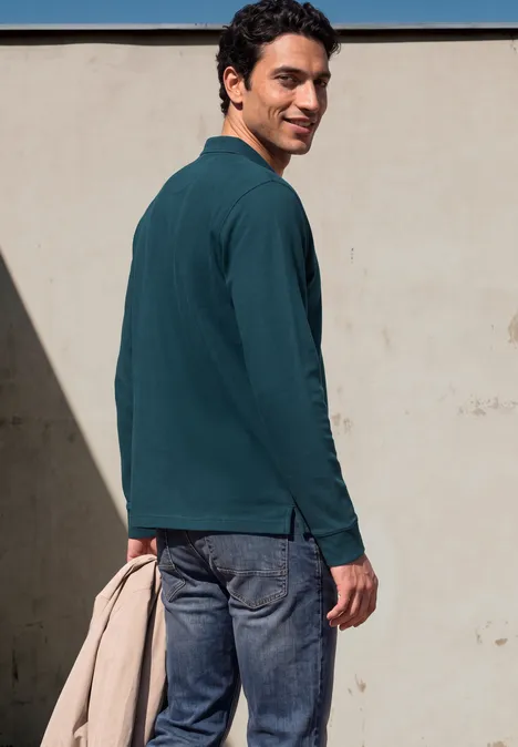Ein Mann in Jeans und grünem Langarm-Poloshirt hält eine beige Jacke, blickt über die Schulter vor einer hellen Wand.