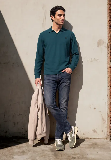 Ein Mann lehnt an einer Wand, trägt grünes Langarmshirt, Jeans, Sneaker und hält eine hellbeige Jacke.