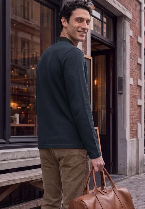 Ein lässig gekleideter Mann verlässt ein Gebäude, trägt eine Lederreisetasche und lächelt zurück.