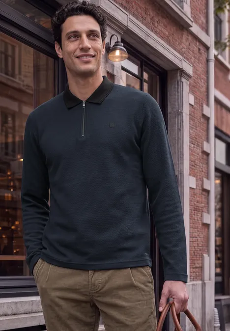 Ein Mann in schwarzem Langarm-Poloshirt und khakifarbenen Hosen mit Tasche vor historischem Gebäude.
