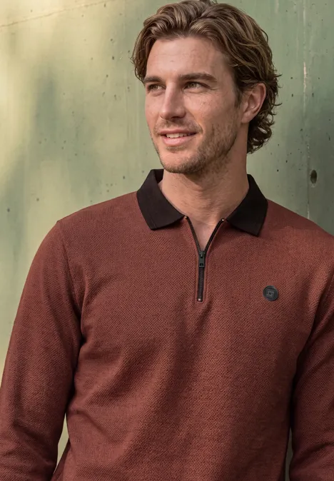 Ein lächelnder Mann in braunem Pullover mit Reißverschluss und schwarzem Kragen vor grünem Metallhintergrund.