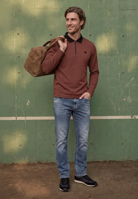 Ein Mann in Jeans und braunem Pullover trägt eine Reisetasche auf der Schulter und steht vor einer grünen Wand.