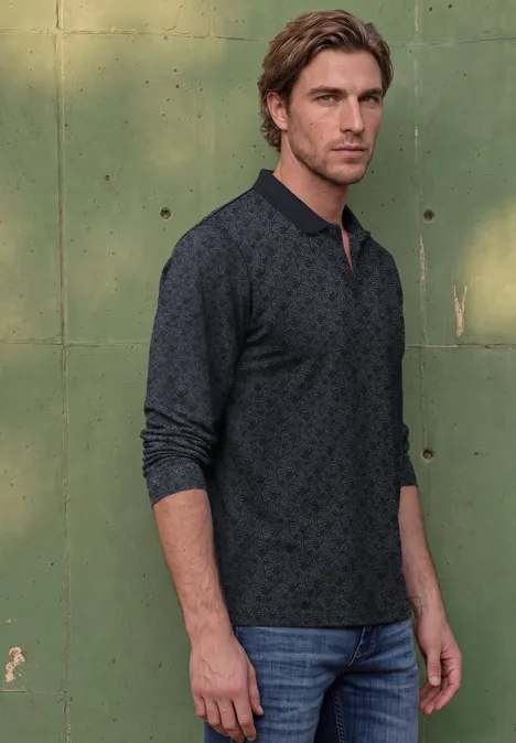 Een man staat voor een groene muur, draagt jeans en een donkergrijs poloshirt met lange mouwen en subtiel patroon.