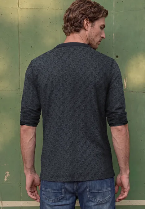 Een man met de rug naar de kijker voor een groene muur, in een donker gemusterd poloshirt en jeans.
