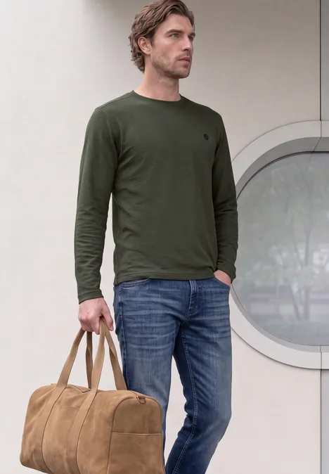 Ein Mann steht in Jeans und olivgrünem Langarmshirt, hält lässig eine braune Reisetasche.