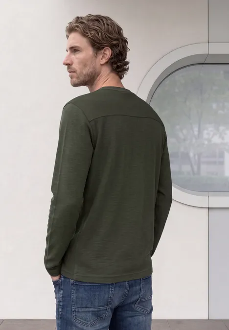 Ein Mann in grünem Langarmshirt und Jeans steht seitlich gewandt vor hellem Hintergrund.