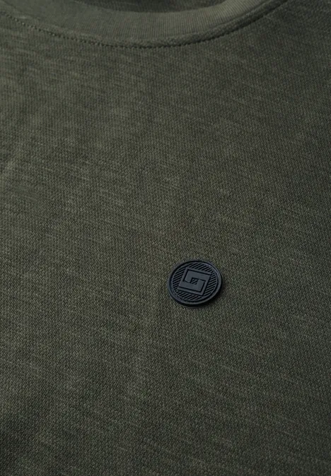 Grünes Strickshirt mit rundem schwarzem Logo-Pin auf der Brust.
