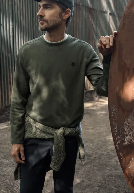 Ein Mann in grünem Sweatshirt und Cap, Pullover um die Hüfte gebunden, steht vor einer Metallstruktur im Freien.