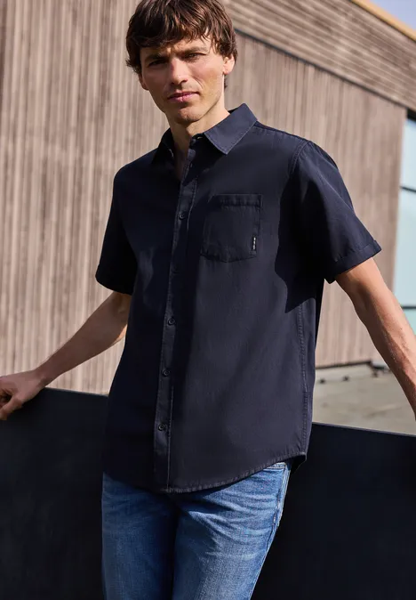 Ein Mann trägt ein schwarzes Kurzarmhemd und blaue Jeans und lehnt lässig an einer Wand.