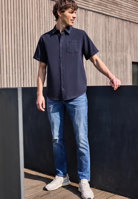 Ein Mann steht vor einer Holzfassade und trägt ein dunkles Hemd, blaue Jeans und weiße Sneaker.