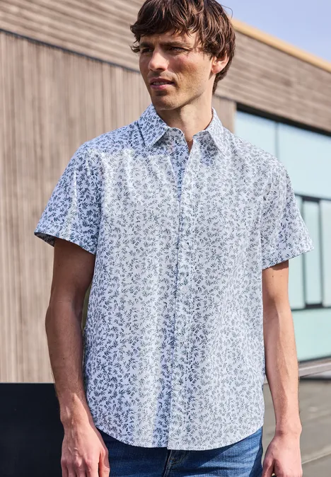 Mężczyzna ma na sobie jasną, wzorzystą koszulę z krótkim rękawem i stoi przed nowoczesnym budynkiem.