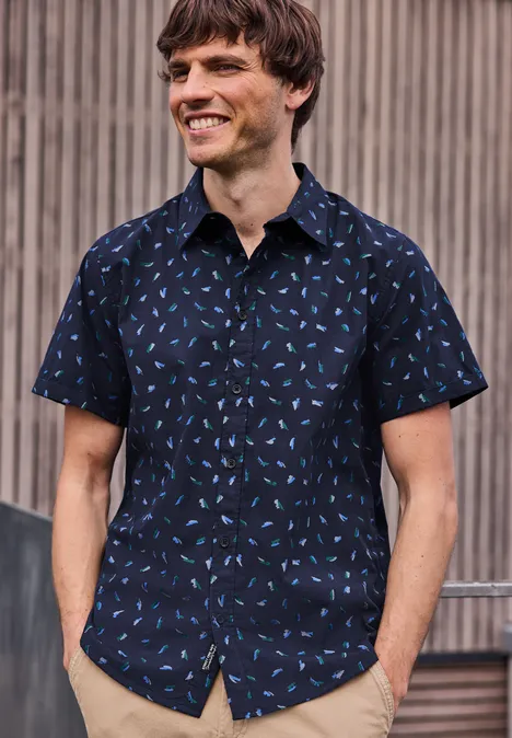 Mężczyzna w granatowej koszuli z jasnym wzorem stoi przed drewnianą ścianą.