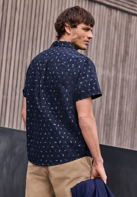 Mężczyzna tyłem stoi przed drewnianą ścianą w granatowej koszuli w wzory i khaki spodniach.