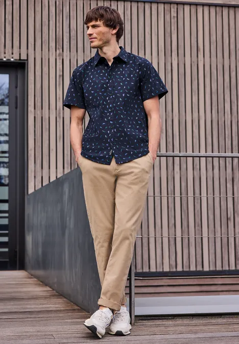 Mężczyzna w wzorzystej koszuli, beżowych spodniach i sneakersach opiera się swobodnie o balustradę na tle drewnianej fasady.