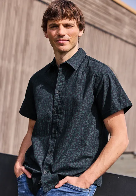 Mężczyzna stoi przy drewnianej ścianie w ciemnoniebieskiej, wzorzystej koszuli z krótkimi rękawami.