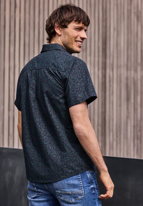 Mężczyzna tyłem w ciemnej koszuli z wzorem i jeansach, patrzy przez ramię na drewniane tło.