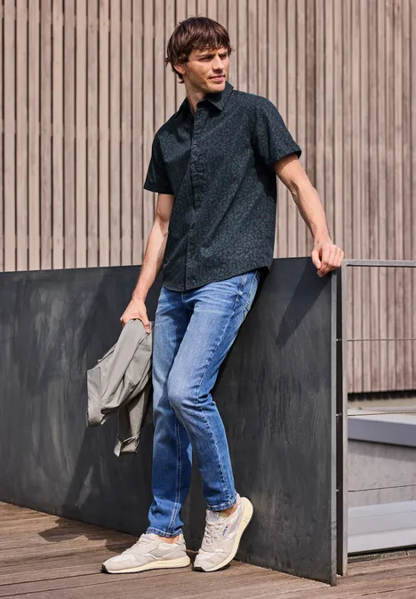 Młody mężczyzna oparty o drewnianą ścianę, w ciemnej koszuli, dżinsach i białych trampkach.