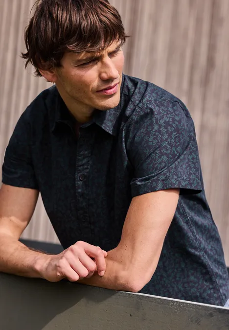 Mężczyzna opiera się o balustradę, ma na sobie ciemną, wzorzystą koszulę z krótkim rękawem na tle drewna.