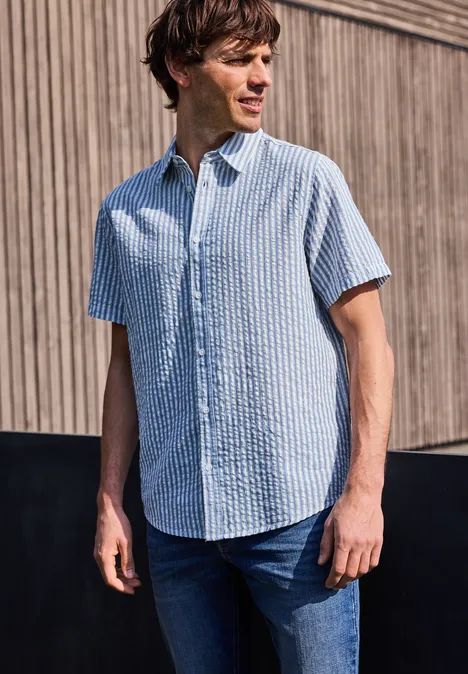 Mężczyzna z rozmazaną twarzą nosi jasnoniebieską krótko rękawową koszulę przed drewnianą ścianą.