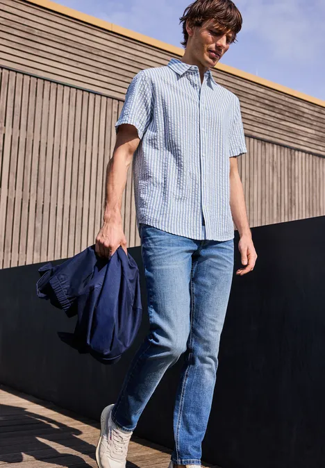 Osoba ubrana w jasnoniebieską koszulę z krótkim rękawem, dżinsy i białe trampki, trzyma kurtkę i stoi na zewnątrz.