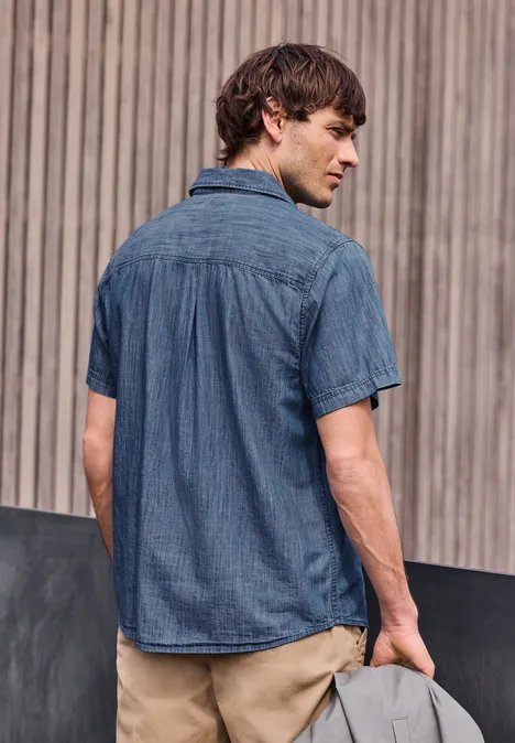 Mężczyzna od tyłu w krótkorękawowej jeansowej koszuli i beżowych chino na tle pionowo strukturalnej ściany.