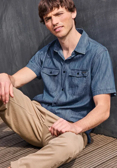 Młody mężczyzna w jeansowej koszuli i beżowych spodniach siedzi na tle ciemnej ściany.