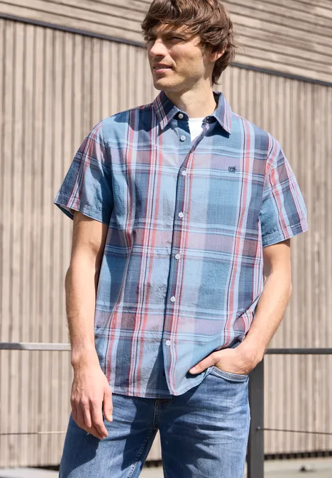 Een man staat voor een houten wand, draagt een geruit overhemd met korte mouwen en jeans, hand in de zak.