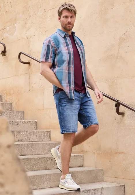 Een man in zomerse kleding staat op een stenen trap en leunt tegen de leuning voor een lichte muur.
