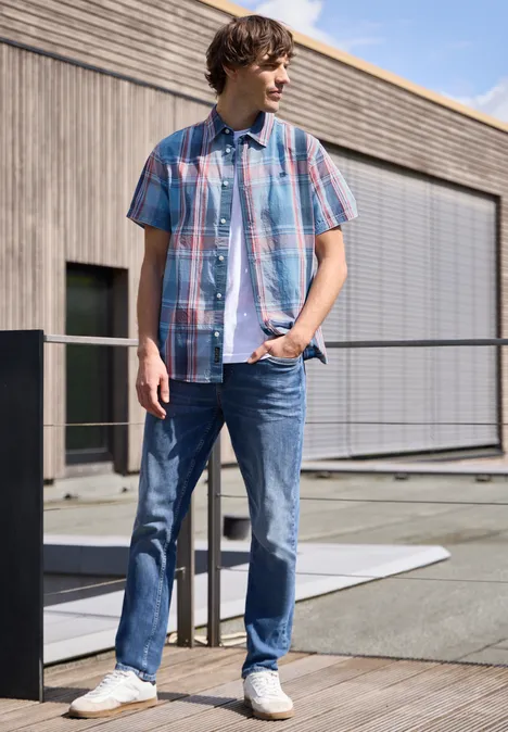 Een jonge man leunt ontspannen op een reling bij een modern gebouw, gekleed in een geruit overhemd en jeans.