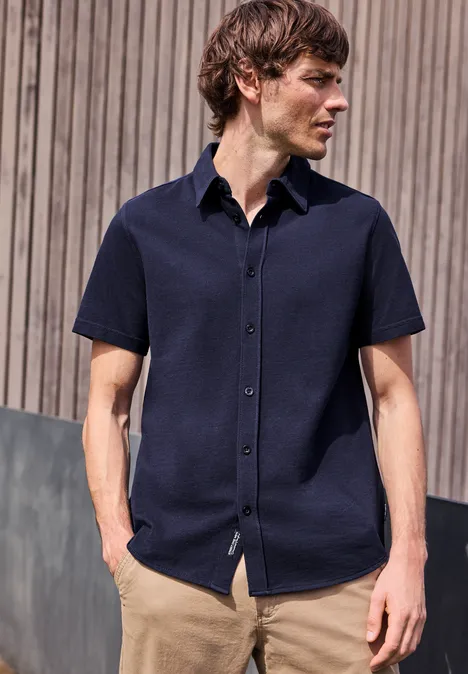 Mężczyzna stoi przed drewnianą ścianą w granatowej koszuli z krótkim rękawem i jasnobeżowych spodniach.