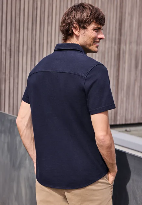 Mężczyzna stoi tyłem do kamery w granatowej koszuli z krótkim rękawem przed drewnianą elewacją.