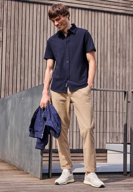 Młody mężczyzna w granatowej koszuli, beżowych spodniach i białych trampkach, trzyma niebieską kurtkę na zewnątrz.