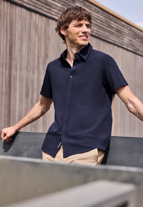 Mężczyzna w granatowej koszuli z krótkim rękawem opiera się o poręcz, za nim drewniana elewacja.