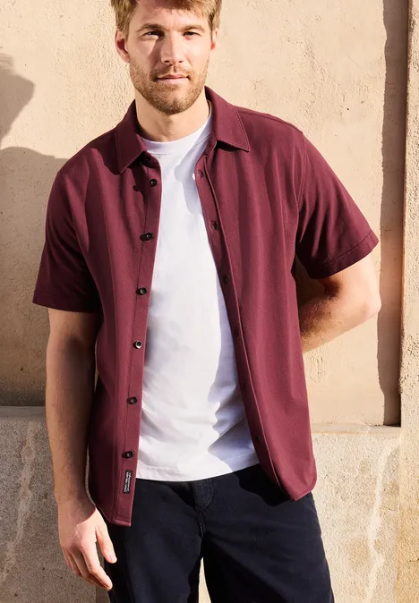 Een man draagt een bordeauxrood overhemd over een wit T-shirt en donkere broek voor een beige muur.