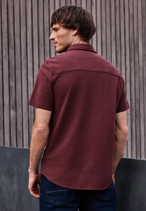 Een man, gezien van achteren, draagt een bordeaux rood overhemd en spijkerbroek voor een houten lattenwand.