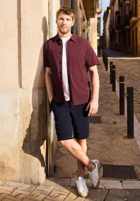 Een man leunt tegen een muur in een zonnig oud straatje, draagt een bordeaux overhemd, short en witte sneakers.