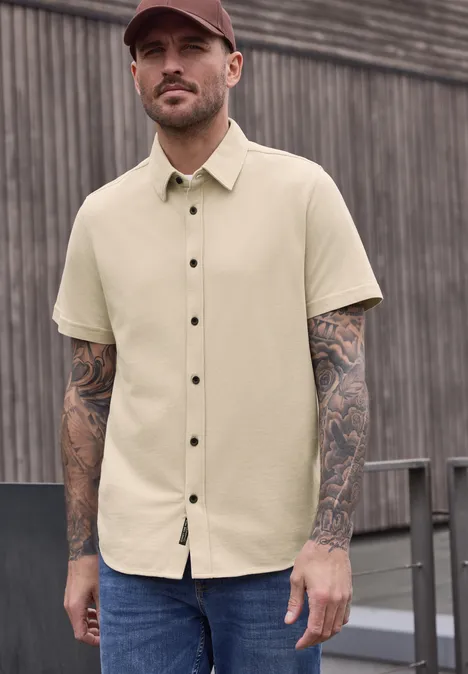 Beiges Kurzarmhemd mit Knöpfen, Tattoos an den Armen, vor einer Holzwand.