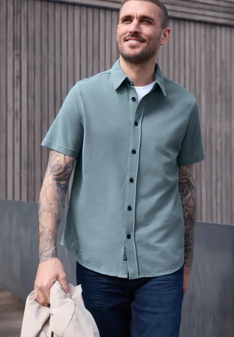 Mann mit grünem Kurzarmhemd, tätowierten Armen und Jeans, hält eine Jacke vor Holzlattenwand.