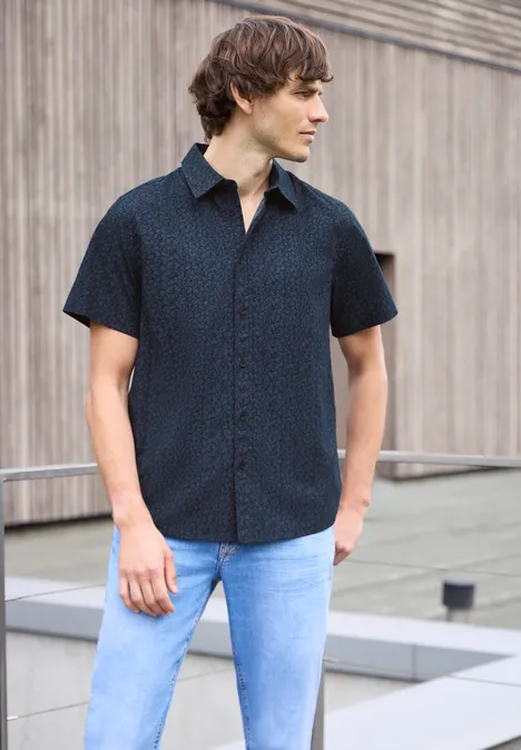 Een man in een donkerblauw overhemd met korte mouwen en lichtblauwe jeans staat voor een houten gevel.