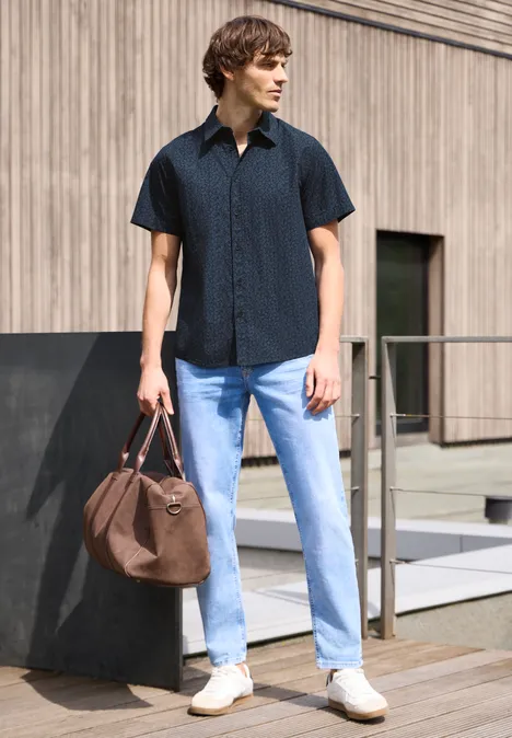 Een man staat voor een modern gebouw, draagt een zwart shirt, lichte jeans, witte sneakers en heeft een bruine tas.